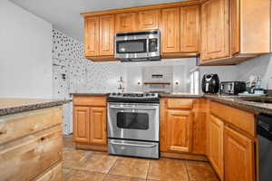 Kitchen with appliances with stainless steel finishes, dark stone counters, light tile patterned floors, and brown cabinetry