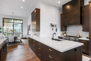 Kitchen with decorative backsplash, dark brown cabinets, hanging light fixtures, ventilation hood, and dark wood-type flooring