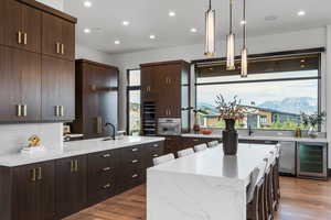 Kitchen with dark brown cabinets, light stone countertops, recessed lighting, wine cooler, and hanging light fixtures