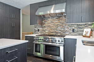 Kitchen with range hood, range with two ovens, backsplash, modern cabinets, and light stone countertops