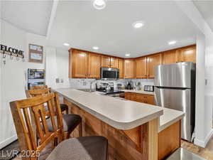 Kitchen featuring a peninsula, appliances with stainless steel finishes, a kitchen bar, brown cabinetry, and light countertops