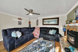 Living room featuring wood finished floors, a textured ceiling, a glass covered fireplace, crown molding, and ceiling fan