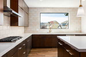 Kitchen with dark brown cabinets, wall chimney range hood, light wood finished floors, stainless steel gas cooktop, and hanging light fixtures