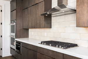 Kitchen featuring wall chimney exhaust hood, appliances with stainless steel finishes, modern cabinets, dark brown cabinetry, and backsplash