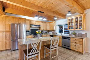 Kitchen with light stone counters, appliances with stainless steel finishes, glass insert cabinets, and a wooden ceiling with exposed beams