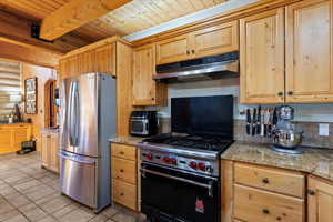 Kitchen featuring stainless steel appliances, under cabinet range hood, light stone counters, a wood ceiling with exposed beams, and light tile patterned floors