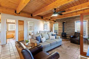 Living room with a wood stove, a wooden ceiling with exposed beams, a ceiling fan, rustic walls, and a wall mounted air conditioner