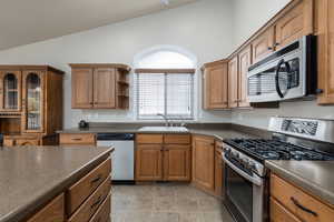 Kitchen featuring appliances with stainless steel finishes, dark countertops, brown cabinetry, open shelves, and vaulted ceiling
