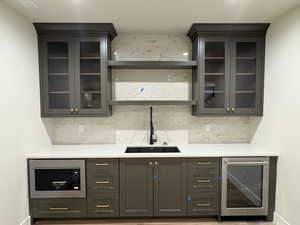 Bar featuring beverage cooler, light stone counters, stainless steel microwave, glass insert cabinets, and backsplash