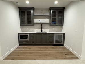 Bar featuring glass insert cabinets, beverage cooler, tasteful backsplash, light wood-type flooring, and stainless steel microwave