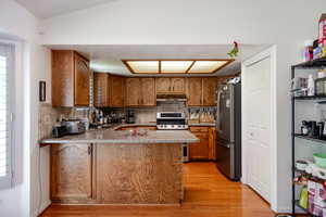 Kitchen with brown cabinetry, decorative backsplash, a peninsula, stainless steel appliances, and light wood-type flooring