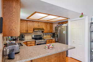 Kitchen featuring a textured ceiling, backsplash, brown cabinets, a peninsula, and appliances with stainless steel finishes