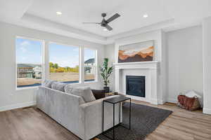 Living area with a tray ceiling, light wood-style floors, a glass covered fireplace, a ceiling fan, and recessed lighting