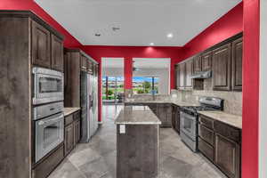 Kitchen featuring dark brown cabinets, appliances with stainless steel finishes, light stone counters, a kitchen island, and recessed lighting
