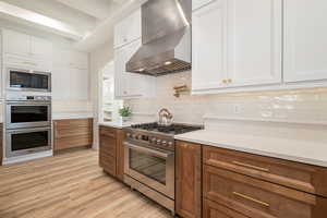 Kitchen with tasteful backsplash, white cabinets, wall chimney exhaust hood, stainless steel appliances, and beam ceiling