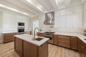 Kitchen featuring brown cabinetry, white cabinetry, beamed ceiling, stainless steel appliances, and wall chimney exhaust hood