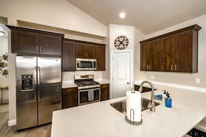 Kitchen with stainless steel appliances, dark brown cabinets, a peninsula, light stone counters, and lofted ceiling