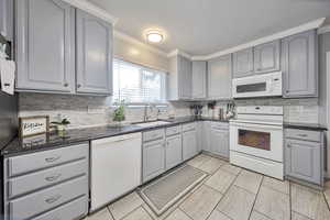 Kitchen with white appliances, gray cabinetry, crown molding, backsplash, and dark stone countertops