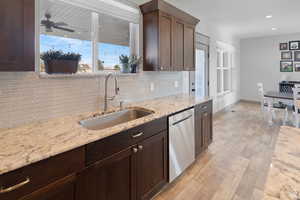 Kitchen with backsplash, light wood-type flooring, light stone countertops, stainless steel dishwasher, and dark brown cabinets