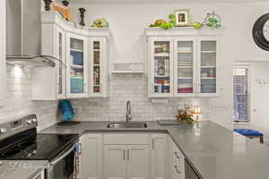 Kitchen featuring stainless steel range with electric stovetop, wall chimney exhaust hood, white cabinetry, glass insert cabinets, and tasteful backsplash
