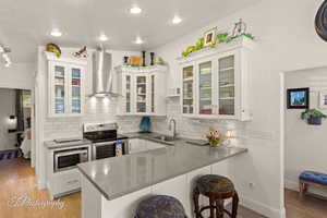 Kitchen featuring a breakfast bar area, light wood finished floors, a peninsula, glass insert cabinets, and wall chimney range hood