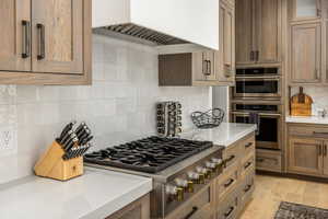 Kitchen featuring decorative backsplash, appliances with stainless steel finishes, custom exhaust hood, light stone countertops, and light wood-style floors