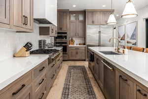 Kitchen featuring pendant lighting, custom exhaust hood, brown cabinetry, appliances with stainless steel finishes, and light stone counters