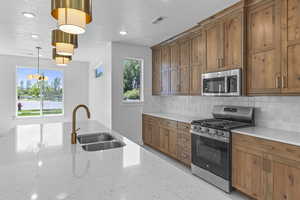 Kitchen with brown cabinetry, stainless steel appliances, decorative backsplash, light stone counters, and pendant lighting