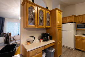 Kitchen featuring freestanding refrigerator, brown cabinetry, glass insert cabinets, light countertops, and stainless steel microwave
