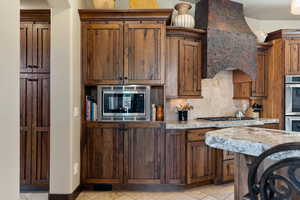 Kitchen featuring decorative backsplash, brown cabinetry, stainless steel appliances, custom exhaust hood, and light stone countertops