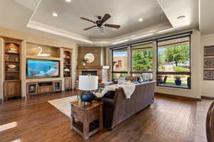 Living area featuring a tray ceiling, dark wood-style floors, a stone fireplace, ceiling fan, and built in features