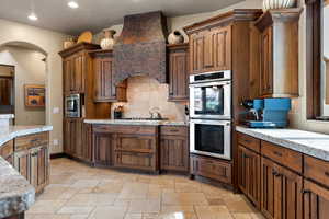 Kitchen with stainless steel appliances, custom exhaust hood, backsplash, stone tile flooring, and recessed lighting