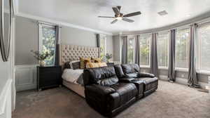 Owner's Suite Bedroom with blackout curtains, bay window with wooded tranquil views, ornamental molding, carpet flooring, a textured ceiling, and a ceiling fan