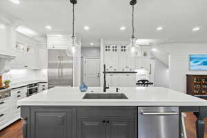 Kitchen with glass insert cabinets, dark wood-style flooring, a center island with sink, white cabinetry, and light stone counters