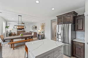 Kitchen with stainless steel fridge, hanging light fixtures, dark brown cabinets, a center island, and light stone counters