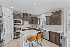 Kitchen with dark brown cabinetry, tasteful backsplash, appliances with stainless steel finishes, light stone countertops, and recessed lighting