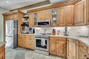 Kitchen featuring glass insert cabinets, stainless steel appliances, light stone countertops, backsplash, and brown cabinets