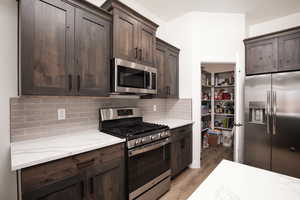 Kitchen with stainless steel appliances, dark brown cabinetry, light wood-type flooring, light stone counters, and backsplash