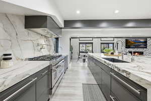 Kitchen with light stone counters, range with two ovens, backsplash, range hood, and a warm lit fireplace