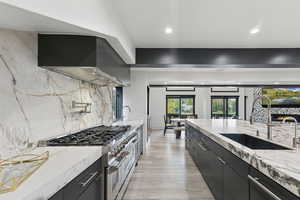 Kitchen with light stone countertops, dark cabinets, range with two ovens, light wood-style floors, and wall chimney range hood