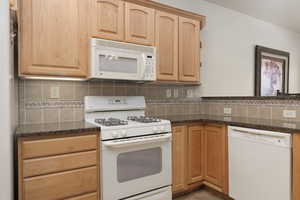 Kitchen featuring white appliances, backsplash, light brown cabinetry, dark stone countertops, and a peninsula