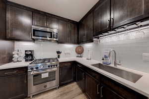 Kitchen featuring dark brown cabinets, stainless steel appliances, tasteful backsplash, and light stone counters
