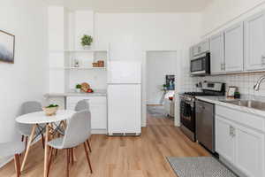 Kitchen with stainless steel appliances, light wood-style floors, open shelves, backsplash, and white cabinetry