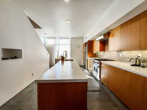 Kitchen featuring an island with sink, stainless steel appliances, light countertops, brown cabinets, and ventilation hood