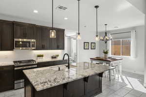 Kitchen with appliances with stainless steel finishes, dark brown cabinets, light stone countertops, hanging light fixtures, and tasteful backsplash