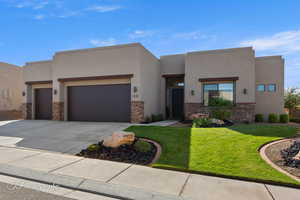 Pueblo-style house featuring a front yard, stone siding, stucco siding, concrete driveway, and a garage