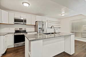 Kitchen with open shelves, stainless steel appliances, light stone countertops, a textured ceiling, and dark wood-style flooring
