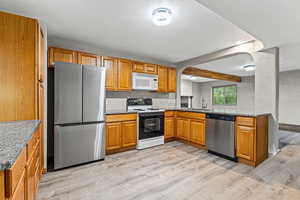 Kitchen with appliances with stainless steel finishes, beam ceiling, light wood-style floors, brown cabinetry, and light stone counters