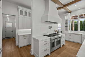 Kitchen with glass insert cabinets, exhaust hood, white cabinets, range with two ovens, and beamed ceiling