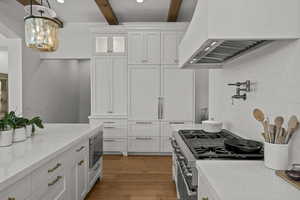 Kitchen featuring white cabinets, custom range hood, light wood finished floors, beamed ceiling, and light stone counters